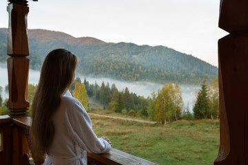 Fototapeta na wymiar Back view of woman in spa bathrobe standing at wooden balcony of hotel resort room and enjoying beautiful morning alpain mountain and forest scenic. No face