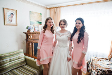 Fabulous young bride posing with two gorgeous bridesmaids in pink dresses in light room.