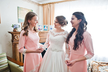 Fabulous young bride posing with two gorgeous bridesmaids in pink dresses in light room.
