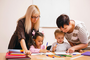 Two teachers in class with their young students with a beautiful young woman helping two small girls at a desk
