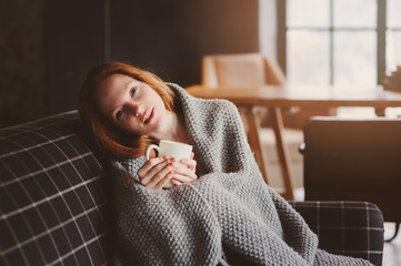 young sick woman healing with hot drink at home on cozy couch, wrapped in knitted blanket
