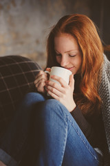 young sick woman healing with hot drink at home on cozy couch, wrapped in knitted blanket