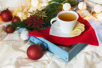 Breakfast with a cup of tea, a biscuit on a tray on the bed on a white blanket in the winter morning decorated with Christmas decorations, spruce branches with luminous garlands, the concept  holiday