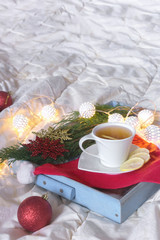 Obraz na płótnie Canvas Breakfast with a cup of tea, a biscuit on a tray on the bed on a white blanket in the winter morning decorated with Christmas decorations, spruce branches with luminous garlands, the concept holiday
