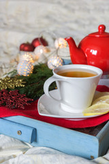 Breakfast with a cup of tea, a biscuit on a tray on the bed on a white blanket in the winter morning decorated with Christmas decorations, spruce branches with luminous garlands, the concept  holiday