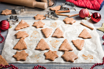 Christmas gingerbread cookies on paper for baking. Fresh baked star and fir-tree shaped biscuits on wooden background.  New Year and Christmas Celebration. Red baubles, berries, garland, ornaments