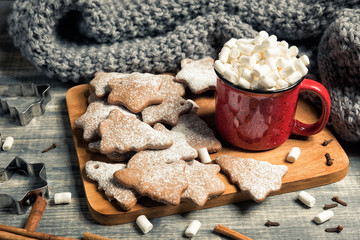 Red cup and gingerbread cookies. Mug of hot chocolate with marshmallow. Fresh baked star and fir-tree shaped biscuits on wooden background. Grey plaid or scarf