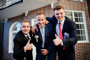 Handsome groom in his wedding tuxedo posing with groomsmen or best men outdoor with brick house on the background.