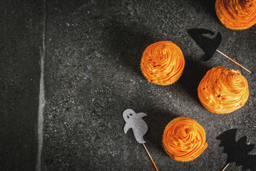 Simple idea of funny kid's treat for Halloween: pumpkin cakes with cream, with decorations in the form of holiday symbols - ghost, witch, bat. On a black background, copy space
