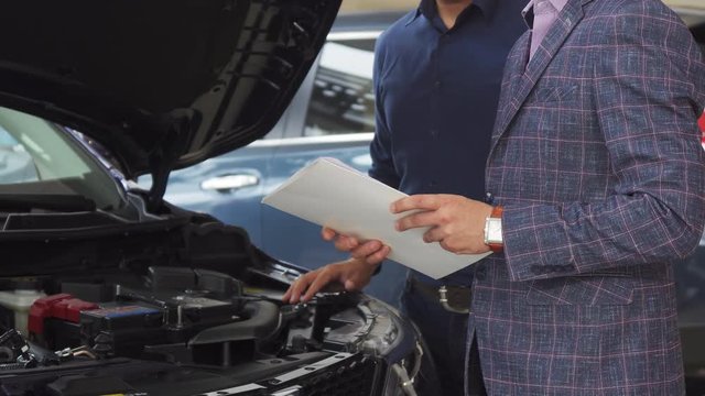 The seller holds a piece of paper with the characteristics of the car. He tells about it to the buyer. He also shows everything in an open hood. Two representative men stand in the car salon