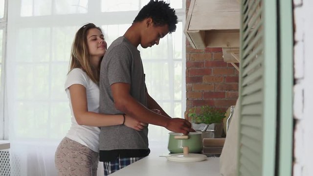 African man preparing food in pot standing in the kitchen. His caucasian girlfriend coming up to him and embracing. Real love. Slowmotion shot