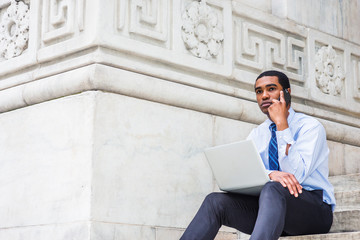 Young African American Businessman working on laptop computer, talking on cell phone, sitting on stairs by marble wall outside office building in New York, wearing white shirt, tie, black pants..