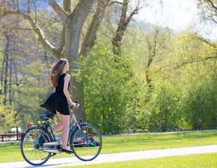 Obraz premium Portrait view of girl on bicycle wearing on black short dress. Young happy Woman riding along road on green spring outdoor Park. Sporty young girl riding a bicycle on a sunny morning
