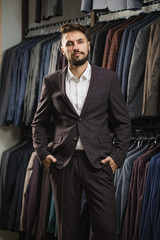 Full length portrait of bearded man in suit near the rack with suits in a shop while looking at the camera