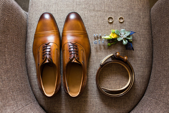 Closeup of elegant stylish brown male accessories isolated on brown textile background of armchair. Top view of leather belt, pair of classic shoes, floral corsage, and two golden wedding rings.