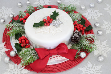 Festive iced christmas cake with red bow, holly, fir, snowflake and bauble decorations with silver foil wrapped chocolates on glitter background.