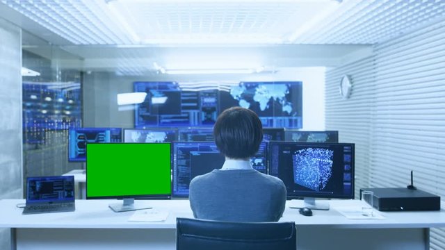 Back View of the IT Technician Working on with Neural Network on His Personal Computer with Green Screen Mock-up. He Works in a Big System Control Data Center with Multiple Monitors Showing Graphics.