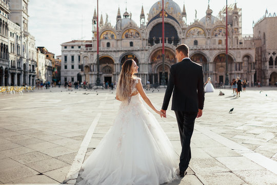 Bride and groom hold their hands together walking on the square in Venice