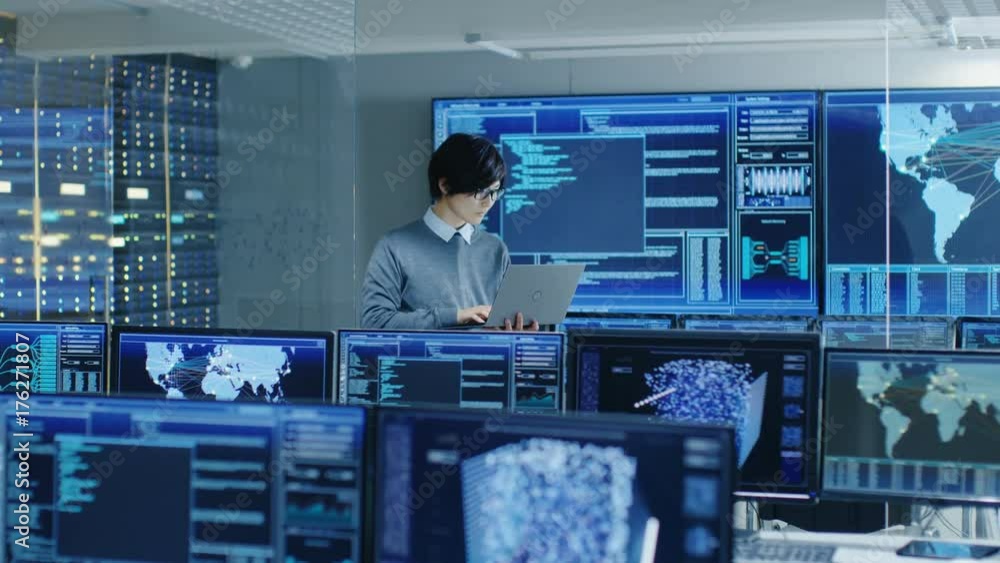 Poster In the System Control Room IT Technician Holds and Works on a Laptop, in Background Multiple Displays with Graphics.  Shot on RED EPIC-W 8K Helium Cinema Camera.