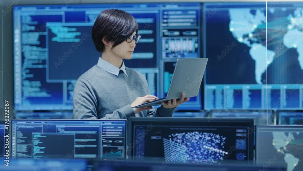 Poster In the System Control Room IT Technician Holds and Works on a Laptop, in Background Multiple Displays with Graphics.  Shot on RED EPIC-W 8K Helium Cinema Camera.