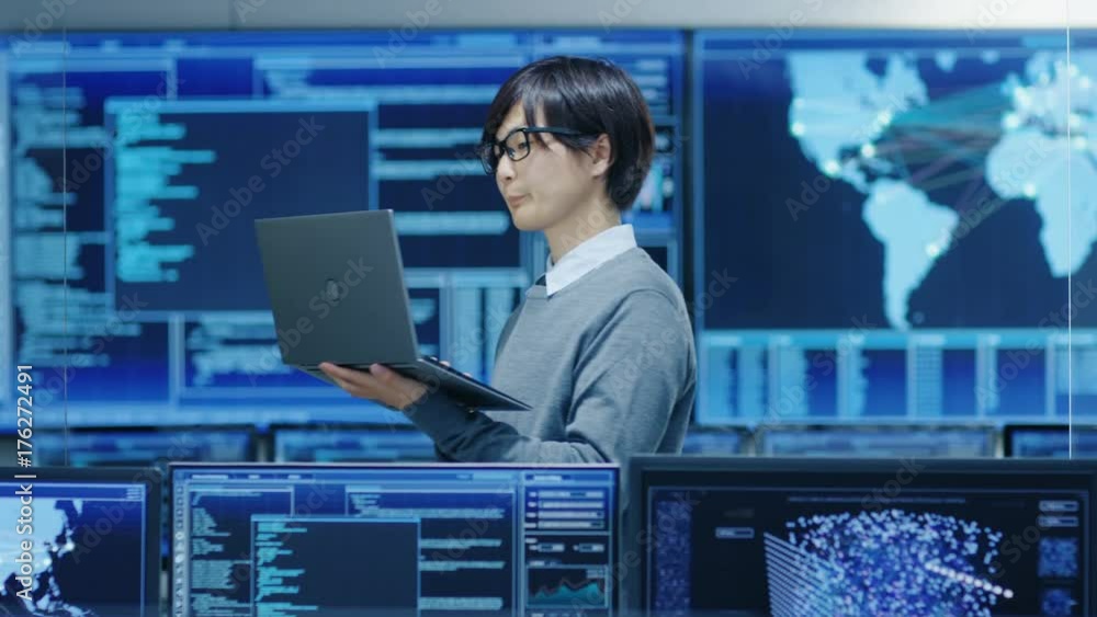 Poster In the System Control Room IT Technician Holds and Works on a Laptop, in Background Multiple Displays with Graphics.  Shot on RED EPIC-W 8K Helium Cinema Camera.