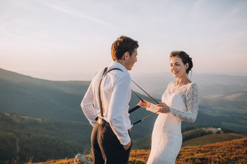Bride plays with groom's braces posing on a golden hill