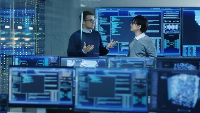 In the Data Center System Control Room Monitors Show Work Done on Neural Networking, AI integration and Data Mining. In the Background Two Specialists Talking.