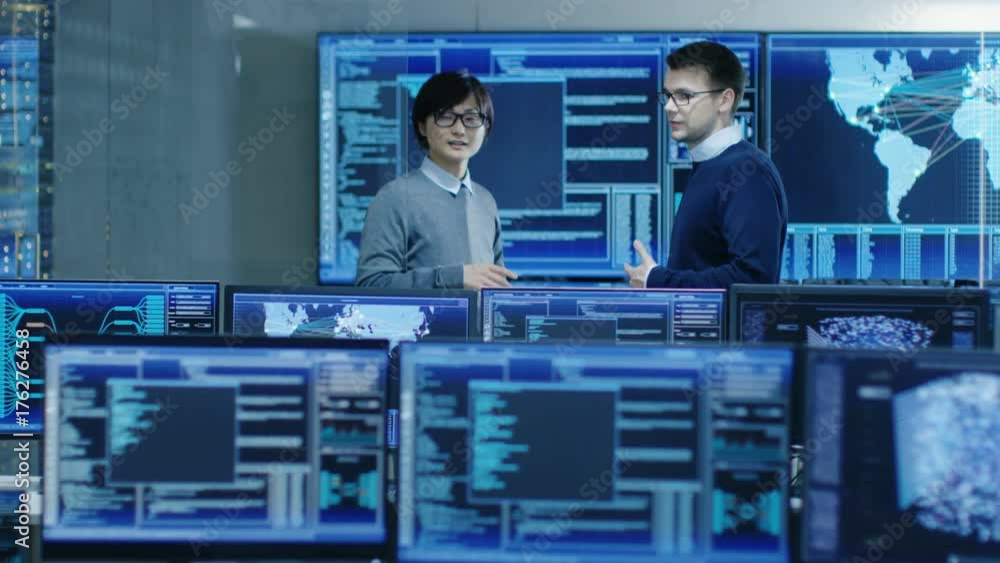 Poster In the System Control Room Project Manage and IT Engineer Have Discussion, they're surrounded by Multiple Monitors with Graphics. They Work in a Data Center on Data Mining, AI and Neural Networking. 