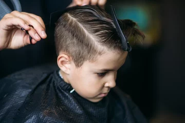 Fotobehang Little Boy Getting Haircut By Barber While Sitting In Chair At Barbershop. © Aleksandr