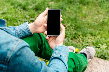 Man use his Mobile Phone outdoor, close up. Mobile phone in hands a young hipster business man in denim shirt and green jeans on the background of green grass.