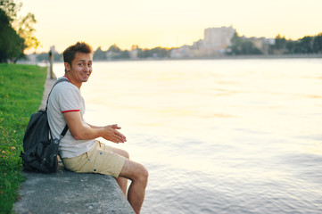 Fototapeta na wymiar Smiling young man sitting on the concrete banks of river, lowering his feet over water, at sunset in summer on blurred background of city panorama.