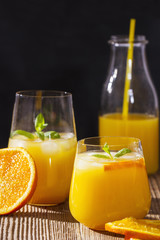 Two glasses of orange juice with ice and mint and a bottle in the background on a wooden table