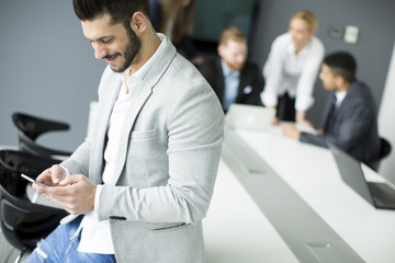 Beard businessman using mobile phone in office while other business people having meeting in background