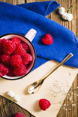 Ripe raspberries in a cup, golden spoon, kitchen towel and flowers on a wooden table, view from top