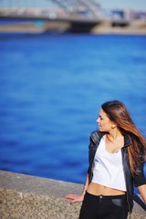 Portrait profile of young beautiful woman with tousled wind long hair into a short t-shirt and leather jacket, standing on the waterfront on background blue waters of Neva.