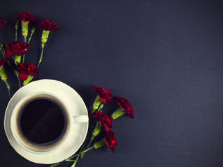A cup of coffee and red carnations on black table, view from top