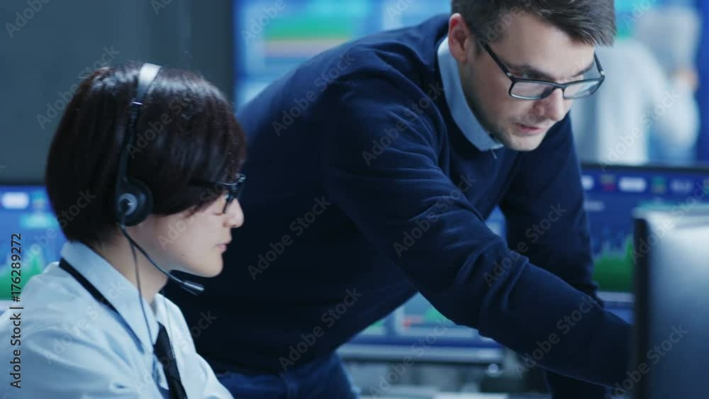 Poster In the Network Operations Center Senior Trader Teaches Young Associate to Makes Personal Client Call with a Headset. They're Surrounded by Monitors Showing Data and Graphs.