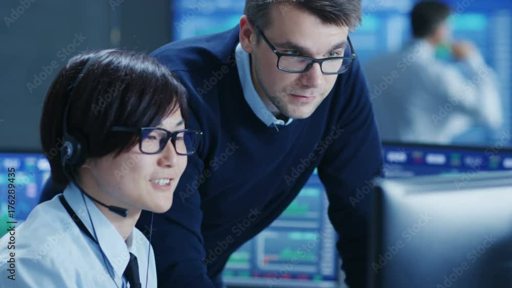 Poster In the Network Operations Center Senior Trader Teaches Young Associate to Makes Personal Client Call with a Headset. They're Surrounded by Monitors Showing Data and Graphs. 
