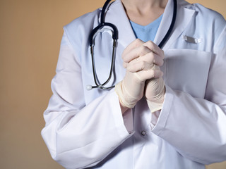 A female doctor in rubber gloves puts his hands together in a fist. Female doctor with a stethoscope gesticulating on a beige background.