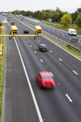 A pair of Average Speed Check cameras above a busy motorway or highway in the UK with speeding traffic below including cars, vans and trucks.