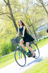 Portrait view of girl on bicycle wearing on black short dress. Young happy Woman riding along road on green spring  outdoor Park. Sporty young girl riding a bicycle on a sunny morning, view from face