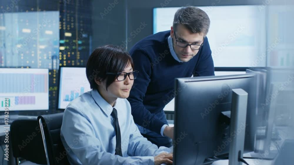 Poster In the Data Mining Center Chief Statistician Consults His Young Assistant sits at His Workstation Surrounded with Monitors Displaying Data and Graphs. Shot on RED EPIC-W 8K Helium Cinema Camera.