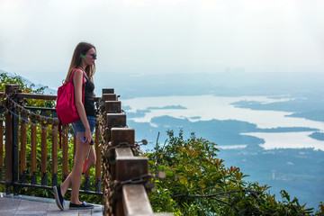 A girl in sunglasses and with a red backpack stands at a fence of a viewing platform. Copy space for text. Walk in the Yanoda National Park, Hainan, China.