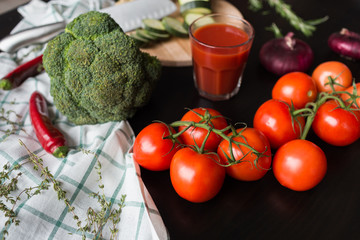 ripe red tomatoes are lying on a black stylish table. in the background we see a glass of tomato juice, red onions, broccoli, red hot chili pepper on a draped fabric and green squash on cutting board