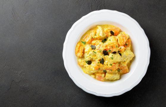 Baked chicken with pumpkin and cream sauce in a white plate on a dark stone background, top view