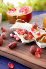 Camembert cheese, orange hard cheese, green grapes, peanuts, walnut and pomegranate seeds on a wooden board on a dark blue background