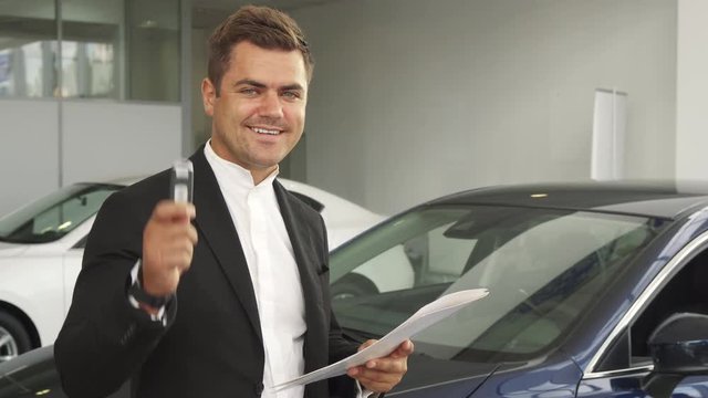 A satisfied buyer reread the contract of buying a car. He shows the keys to his new car. He smiles. Men are in the car showroom