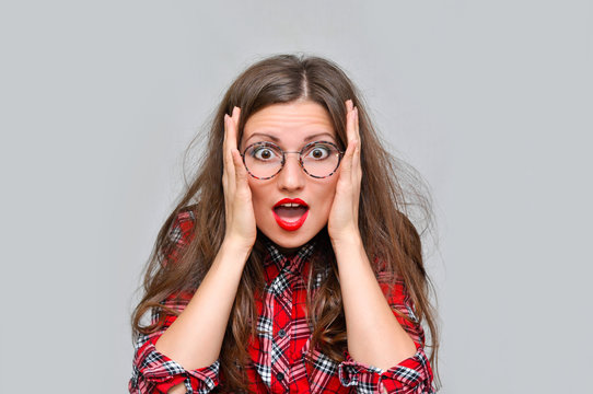 shocked woman in a red checkered shirt and glasses holding her head in both hands
