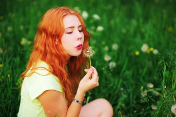 Wonderful portrait of a beautiful redheaded woman in a clearing among the flowers. Girl holding dandelion and blows him.