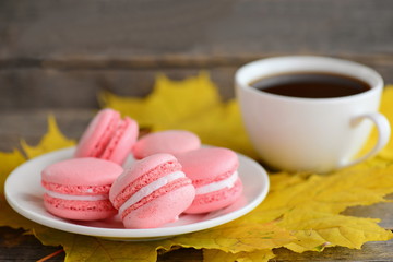 Delicious French macarons. Light pink macarons on a white plate, a cup of coffee, yellow leaves on an old wooden background. Fall breakfast concept. Closeup
